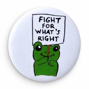 May include: A white button with a green frog holding a sign that reads "FIGHT FOR WHAT'S RIGHT" in black text. The frog has large black eyes and is smiling. The button has a glossy finish.