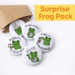 Puede incluir: Un conjunto de botones blancos con ilustraciones de ranas verdes y varias frases, incluyendo "Fight for What's Right" y "Vote". Los botones están cerca de una bolsa de papel marrón, con el texto "Surprise Frog Pack".