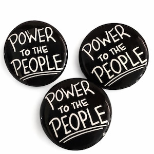May include: Three black round buttons with the white text "POWER TO THE PEOPLE". The text is in a handwritten style. The buttons are arranged in a triangular pattern against a white background.