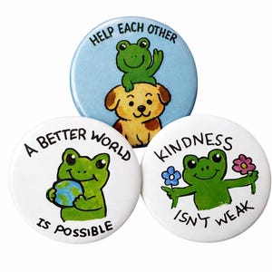 May include: Three round pinback buttons with illustrations and text. One button shows a frog on a dog with the text "Help Each Other." Another shows a frog holding a globe with the text "A Better World Is Possible." The last shows a frog with flowers and the text "Kindness Isn't Weak."