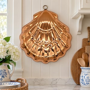 May include: A large, polished copper mold shaped like a scallop shell with a circular hanging loop at the top. The surface features deep, radiating ridges and a reflective metallic finish, designed for decorative wall display or culinary use.
