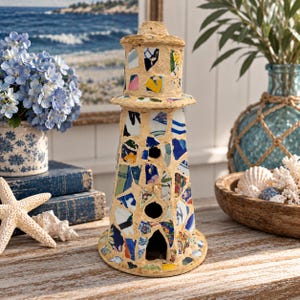 May include: A decorative lighthouse sculpture crafted from a mosaic of colorful ceramic shards embedded in a light-colored mortar base. The structure features a conical shape with a lantern room at the top and several small arched openings.