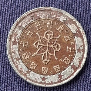 Könnte beinhalten: Eine abgenutzte 2-Euro-Münze in Kupferfarbe. Die Münze zeigt ein zentrales Blumendesign, umgeben von Text und Symbolen. Der äußere Rand ist silberfarben, mit Sternen und weiterem Text. Die Münze weist Alters- und Gebrauchsspuren auf.