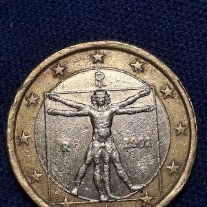 May include: A one euro coin featuring the Vitruvian Man, a famous drawing by Leonardo da Vinci, within a square. The coin has a gold outer ring with stars and the year 2002. The background is a dark blue.