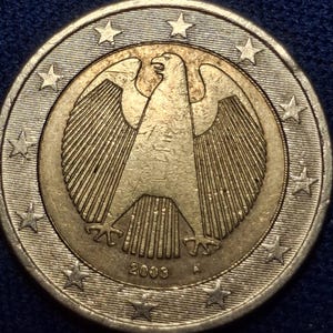 Peut inclure: Une pièce de deux euros de 2003 avec un aigle allemand. La pièce a un centre doré avec l'aigle et l'année, entouré d'un anneau extérieur argenté avec des étoiles. La pièce est posée sur une surface bleu foncé.