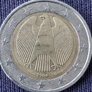 Può includere: Una moneta da due euro d'argento e oro con l'emblema dell'aquila tedesca. La moneta presenta l'anno 2004. L'anello esterno ha delle stelle. La moneta è su una superficie di tessuto blu.