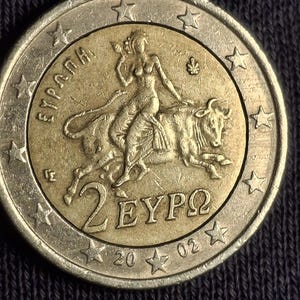 Puede incluir: Una moneda de dos euros con un centro dorado y un anillo exterior plateado. El centro dorado presenta una mujer montando un toro, con el texto "2 EYPΩ" y el año "2002". El anillo exterior tiene estrellas y la palabra "ΕΥΡΩΠΑ".