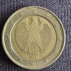 Puede incluir: Una moneda de dos euros de 2002, con un emblema de águila detallado en el centro. La moneda tiene un anillo exterior plateado con estrellas y un círculo interior dorado. El año 2002 está inscrito en la parte inferior.