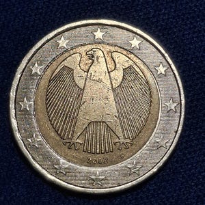 Può includere: Una moneta da due euro raffigurante un'aquila tedesca dettagliata con le ali spiegate, circondata da stelle. La moneta ha un anello esterno argentato e un centro dorato, con l'anno 2002 visibile.