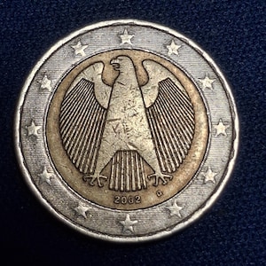 May include: A two-euro coin with a silver outer ring and a gold-coloured centre. The centre features a detailed eagle emblem, with the year 2002 below. The coin's edge is lined with stars.