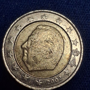 Könnte beinhalten: Eine Zwei-Euro-Münze mit goldenem Zentrum und silbernem Außenring. Die Münze zeigt das Profil eines Mannes, Sterne und das Jahr 2002. Die Münze hat eine Krone und den Buchstaben A. Die Münze liegt auf einer dunkelblauen Oberfläche.