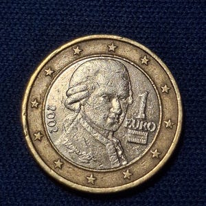 May include: A one euro coin with a portrait of a man in a powdered wig. The coin has a gold outer ring with stars and the year 2002. The centre of the coin is silver-coloured and has the number 1 and the word EURO.