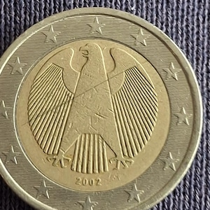 May include: A two-euro coin with the German eagle design. The coin has a gold centre with the eagle and the year 2002, encircled by a silver ring with stars. The coin is on a textured blue surface.