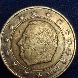 Può includere: Una moneta da due euro del 2003 di colore oro. La moneta presenta il profilo di un uomo, circondato da stelle. L'anno 2003 è visibile nella parte inferiore della moneta. La moneta ha un anello esterno argentato.