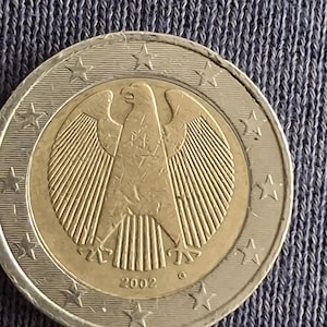Puede incluir: Una moneda de dos euros con un águila alemana detallada, fechada en 2002. La moneda tiene un anillo exterior plateado con estrellas y un centro dorado. El águila es el elemento central del diseño.