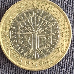 Può includere: Una moneta da un euro del 1999, con un disegno ad albero e le parole "EGALITE FRATERNITE". La moneta ha un anello esterno dorato e un cerchio interno argentato. L'anno 1999 è visibile. Il diametro della moneta è di circa 2,3 cm.