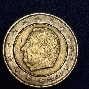 Może przedstawiać: Złota moneta z profilem głowy, otoczona gwiazdami. Na monecie widnieje rok 2000. Moneta jest umieszczona na ciemnoniebieskim tle.