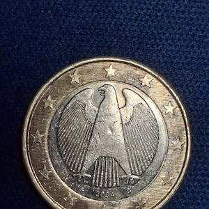 Könnte beinhalten: Eine Ein-Euro-Münze mit einem detaillierten Adler-Emblem mit ausgebreiteten Flügeln, umgeben von Sternen. Die Münze hat einen silbernen Kern und einen goldfarbenen Außenring. Das Jahr 2004 ist auf der Münze zu sehen.