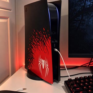 May include: A black and red gaming console featuring a white spider emblem and a black web-like graphic pattern on the side panel. The console is standing vertically on a circular base with a white charging cable plugged into the front port.