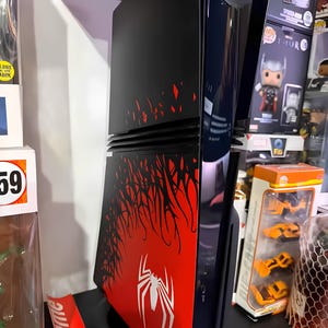 May include: Black and red PlayStation 5 console with a Spider-Man design. The console features a red panel with a white spider logo and a black panel with red accents. The console is displayed vertically.