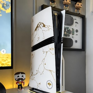 Op de afbeelding: Een witte en zwarte gameconsole met een gouden berg- en kaartontwerp. De console heeft een zwarte horizontale streep en gouden rand. De basis van de console heeft een geel licht. Een klein beeldje staat op de tafel.