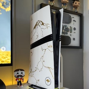 Może przedstawiać: Biało-czarna konsola do gier ze złotym wzorem góry i mapy. Konsola ma czarny pasek pośrodku i świecącą zieloną podstawę. Mała figurka stoi po lewej stronie konsoli.