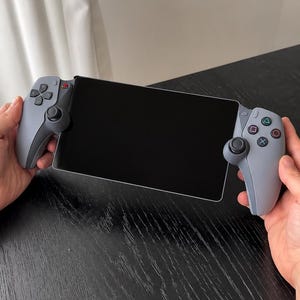 Pode incluir: Um dispositivo de jogo portátil com um ecrã preto retangular central ladeado por dois punhos de comando cinzentos claros. O punho esquerdo tem um direcional e um analógico, enquanto o direito inclui quatro botões de ação e um analógico.