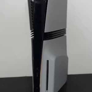 May include: A gray and black gaming console, standing upright. The console has a sleek, modern design with a curved shape. The front panel is gray, while the sides and top are black. A vertical vent is visible on the side.