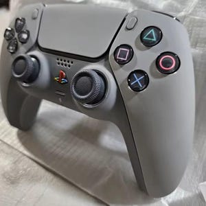 May include: A grey wireless games controller with black joysticks and coloured action buttons. The controller has a touchpad and a PlayStation logo. The buttons are coloured blue, pink, green, and purple.