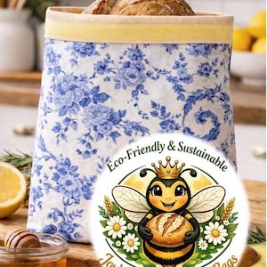 Puede incluir: Una bolsa de pan reutilizable con un estampado floral azul y un ribete amarillo, llena de una hogaza de pan. La bolsa presenta un logotipo circular con una abeja de dibujos animados sosteniendo pan, y el texto "Eco-Friendly & Sustainable" y "Joy's Happy Beeswax Bags".
