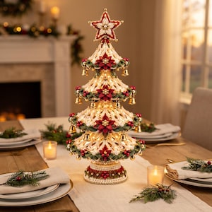 Puede incluir: Un árbol de Navidad decorativo de mesa hecho con encaje de ganchillo color crema, adornado con flores de pascua rojas, pequeñas campanas doradas y bayas rojas. El árbol está coronado con una estrella y tiene una base iluminada con borde rojo y verde.