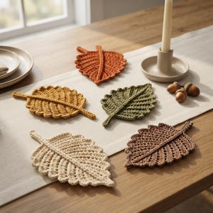 May include: Five crochet leaf coasters in various colours including orange, yellow, olive green, cream, and brown. Each coaster features a textured, ribbed design with a central stem and serrated edges, arranged on a light-coloured table runner.