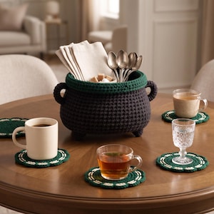 May include: A dark charcoal crochet cauldron basket with a forest green rim and two side handles, containing beige napkins, silver spoons, and sugar cubes. Four round dark green crochet coasters with white stitched edges sit on the table.