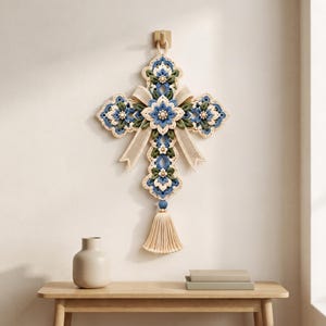 May include: A decorative wall hanging in the shape of a cross, crafted from cream, blue, and sage green yarn. The design features intricate floral crochet patterns, a central beige ribbon bow, and a long cream-colored tassel at the bottom.