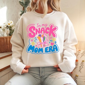 Puede incluir: Sudadera color crema con el texto "In my Snack Mom Era" en rosa y azul. El diseño incluye un manojo de palitos de aperitivo coloridos atados con una cinta rosa, estrellas y corazones.