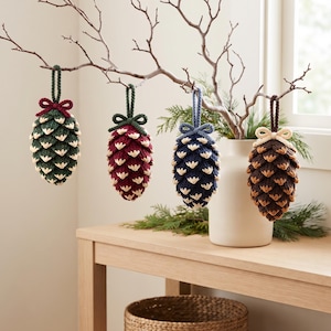 May include: Four crocheted pinecone ornaments hanging from a branch. Each ornament features a unique color combination: dark green with a maroon bow, maroon with a dark green bow, navy blue with a blue bow, and brown with a cream-colored bow.
