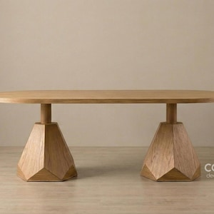 Puede incluir: Una mesa de centro de madera marrón claro con una tapa ovalada. La mesa tiene dos bases de pedestal geométricas y facetadas. La mesa está hecha de madera de color claro y está sobre un fondo neutro. La mesa mide unos 152 cm de largo.