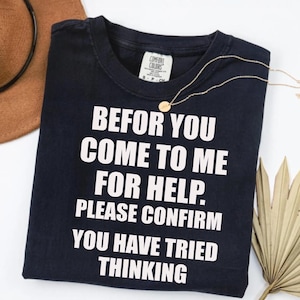Peut inclure: T-shirt bleu marine avec l'inscription blanche : "BEFOR YOU COME TO ME FOR HELP. PLEASE CONFIRM YOU HAVE TRIED THINKING". Un collier doré et un chapeau marron sont également visibles.