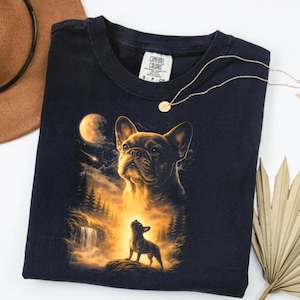 May include: Dark navy blue t-shirt featuring a detailed illustration of a French Bulldog with a forest and moon backdrop. The design includes a waterfall and a smaller dog silhouette. A gold necklace and a brown hat are also visible.