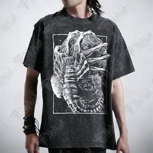 Könnte beinhalten: Dunkelgraues T-Shirt mit einer großen, detaillierten weißen Grafik eines Kreaturenkopfes und -oberkörpers. Das Design ist in einem weißen rechteckigen Rahmen platziert. Das Shirt hat ein Vintage-Aussehen.