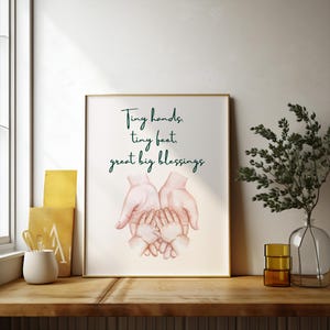 Peut inclure: Une affiche encadrée avec le texte "Tiny hands, tiny feet, great big blessings" en vert. L'œuvre présente une illustration à l'aquarelle de mains d'adultes berçant de petites mains. Le cadre est doré.