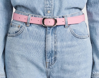 Puede incluir: Un cinturón de cuero rosa claro con acabado liso y una hebilla ovalada de tono plateado pulido. El cinturón está colocado en las trabillas de unos vaqueros de mezclilla azul claro, combinados con una camisa de mezclilla azul claro a juego.
