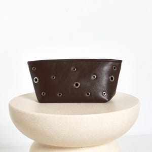 Puede incluir: Un estuche de cuero marrón oscuro con múltiples ojales plateados. La bolsa rectangular se exhibe sobre un pedestal color crema. La bolsa es de color marrón oscuro y tiene un diseño sencillo.