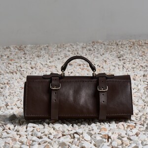 Puede incluir: Un bolso enrollable de cuero marrón oscuro con asa y dos correas con hebilla. El bolso rectangular parece estar hecho de cuero de alta calidad. El bolso está sobre un lecho de guijarros blancos y grises.