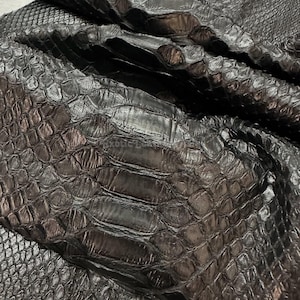 May include: Close-up of black exotic leather with a textured, scale-like pattern. The leather is folded, revealing the natural grain and variations in the material. The surface reflects light, highlighting the dark color and unique texture.