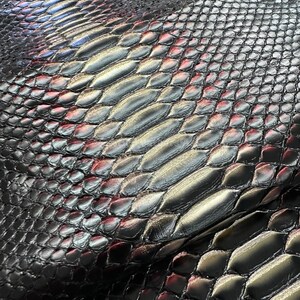 May include: Close-up of a textured, dark gray material with a scale-like pattern. The scales have a metallic sheen, with hints of red and silver, creating a reptile skin appearance. The material appears to be leather.
