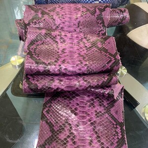 Könnte beinhalten: Nahaufnahme von mehreren Stücken rosa und schwarzem Schlangenleder. Das Leder hat ein natürliches Muster und eine natürliche Textur mit einer Mischung aus rosa und schwarzen Schuppen. Die Stücke sind übereinander gestapelt.