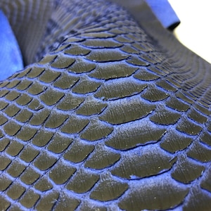 Può includere: Primo piano di un materiale blu scuro strutturato con un motivo a scaglie. Le scaglie sono delineate in un blu più chiaro, creando un contrasto. Il materiale sembra essere pelle o un tessuto simile.