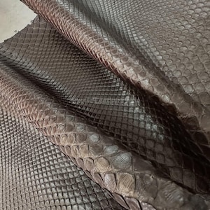 May include: Close-up of a sheet of brown snakeskin leather. The leather has a textured, scaly pattern with variations in color, ranging from dark brown to lighter shades. The material is folded, revealing the underside and adding depth to the image.