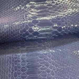 Puede incluir: Primer plano de una lámina de cuero de serpiente de color lavanda. El cuero tiene un patrón de escamas con un acabado brillante. Las escamas son una mezcla de pequeñas y grandes, creando una superficie texturizada. La imagen está bien iluminada.
